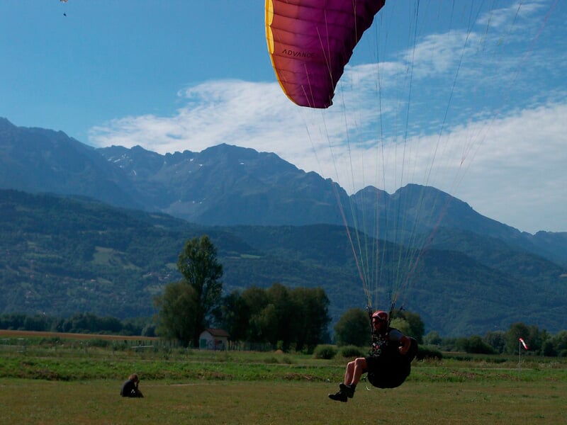 Parapentiste en vol proche du sol avec voile jaune et rose, montagnes en arrière-plan et ciel bleu clair.