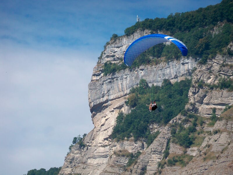 Parapentiste en vol près d'une falaise rocheuse avec voile bleue, ciel clair et végétation dense sur la falaise.