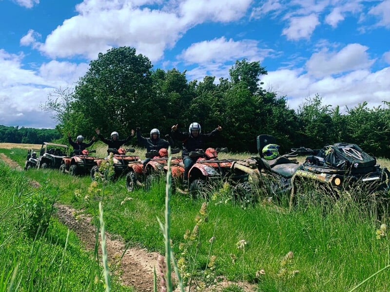 Six quads boueux alignés sur un chemin vert, leurs conducteurs en combinaison noire et casque, entourés de végétation et ciel bleu partiellement nuageux.