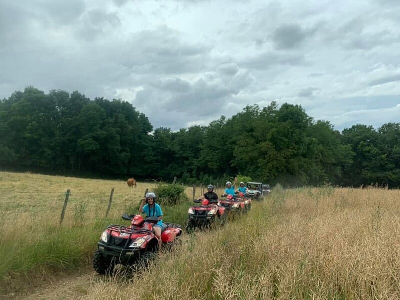 Groupe de personnes en file sur des quads rouges circulant sur un sentier herbeux entre clôture et forêt, sous un ciel nuageux.