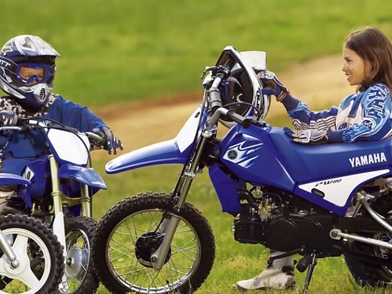 Deux enfants en casque discutent près de motos bleues Yamaha sur un terrain de pilotage en herbe.
