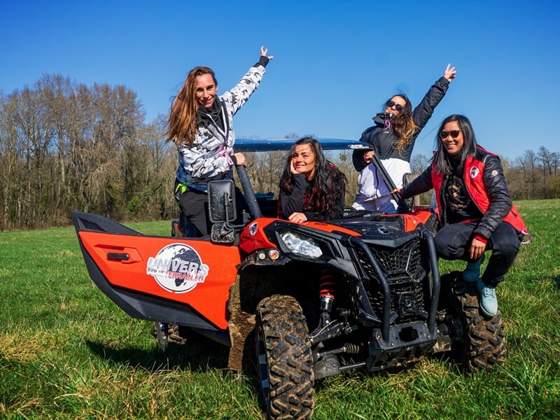 Quatre femmes souriantes posent autour d’un buggy rouge sur une prairie, bras levés sous un ciel bleu dégagé, avec forêt en arrière-plan.