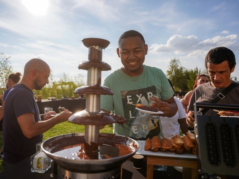 Homme souriant devant une fontaine de chocolat en plein air, tenant une assiette avec des gaufres, entouré de personnes dégustant des gourmandises sous un ciel bleu.