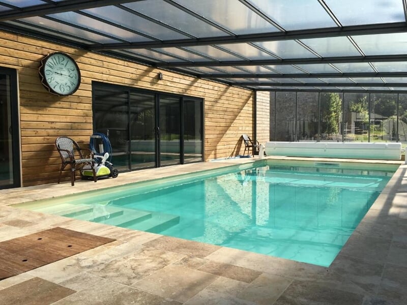 Piscine intérieure avec eau turquoise, entourée de dalles beiges, murs en bois clair, grande horloge murale et verrière laissant entrer la lumière naturelle.