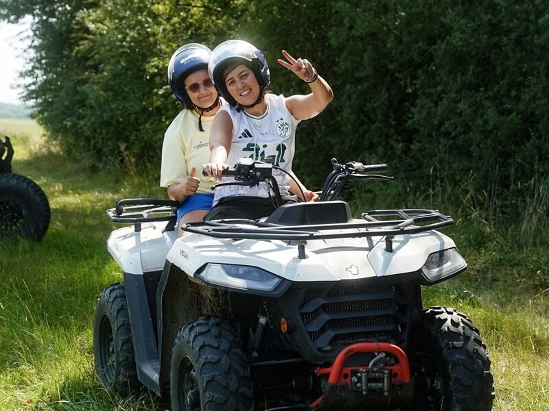 Deux personnes souriantes portant un casque font une randonnée en quad sur un chemin herbeux entouré d'arbres verts.
