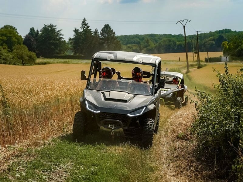 Deux quads tout-terrain traversent un chemin étroit bordé de champs de blé sous un ciel partiellement nuageux.