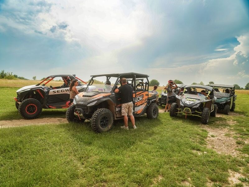 Groupe de personnes autour de plusieurs quads garés sur une prairie verte sous un ciel bleu avec quelques nuages.
