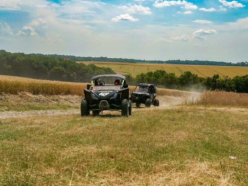 Deux quads tout-terrain roulent sur un chemin de campagne poussiéreux entouré de champs et sous un ciel clair.