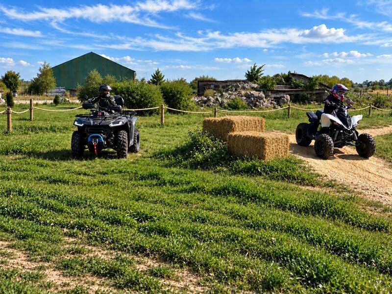 Deux personnes conduisent des quads sur un chemin de terre entouré d'herbe verte et de bottes de paille.