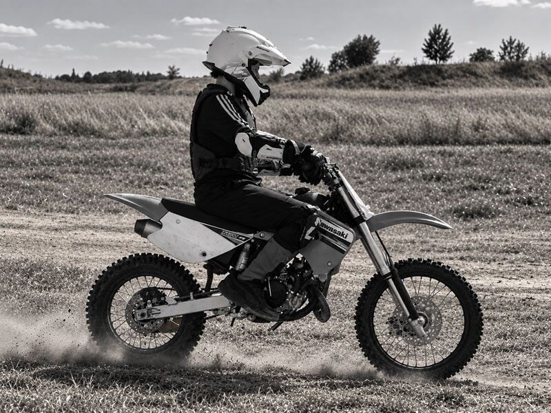Un pilote en tenue complète roule sur une moto tout-terrain sur un terrain herbeux, photo en noir et blanc.