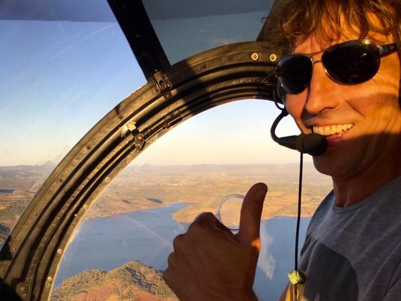 Homme souriant donnant un pouce en l'air à l'intérieur d'un hélicoptère, avec une vue sur un lac à travers la fenêtre.