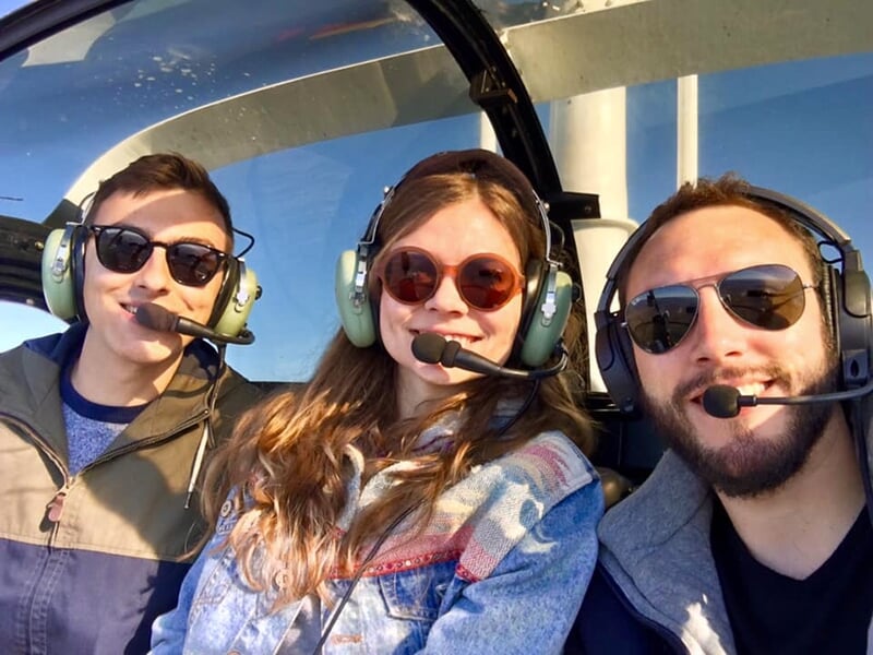 Trois personnes dans l'habitacle d'un hélicoptère, portant des lunettes de soleil et des casques avec micro.