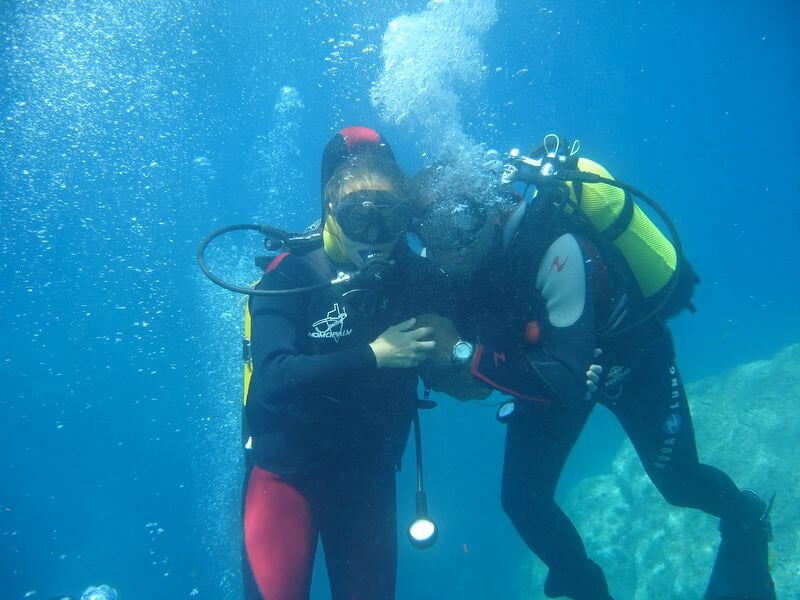 Deux plongeurs sous l'eau en combinaison complète, l'un aidant l'autre lors d'une initiation.
