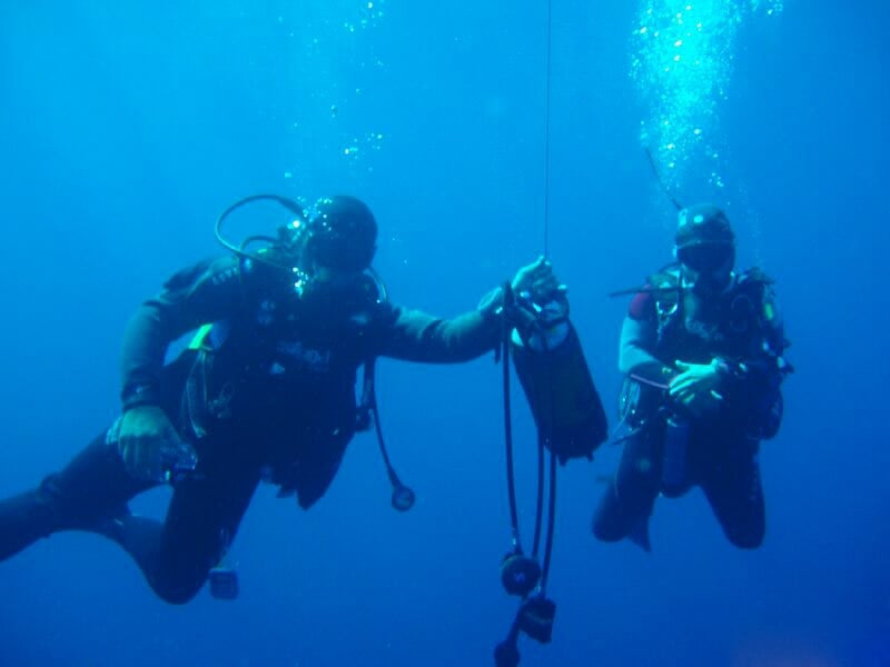 Deux plongeurs sous-marins tenant un équipement, évoluant dans une eau bleue profonde.