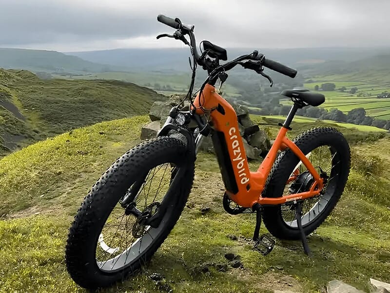Un fat bike orange de la marque Crazybird posé sur une colline verdoyante avec une vue panoramique sur la vallée.