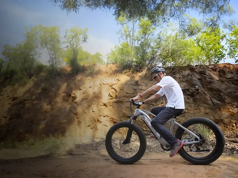 Un cycliste portant un casque roule en fat bike blanc sur un sentier terreux en pleine nature.
