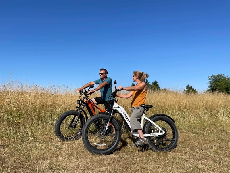 Un couple en fat bike, l’un orange et l’autre blanc, s’arrêtent dans une prairie dorée sous un ciel bleu clair.