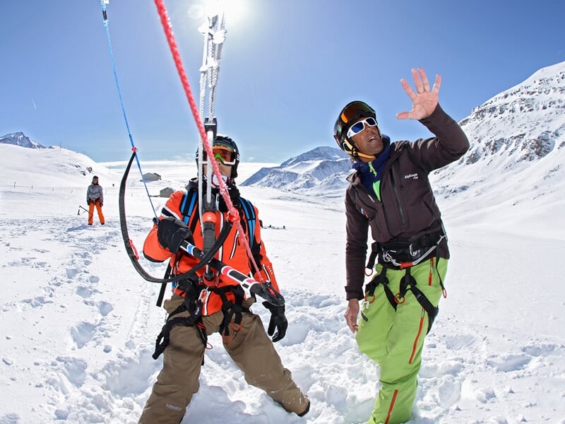 Deux personnes en tenue de neige discutent et préparent leur équipement de snowkite sur un terrain enneigé en montagne.