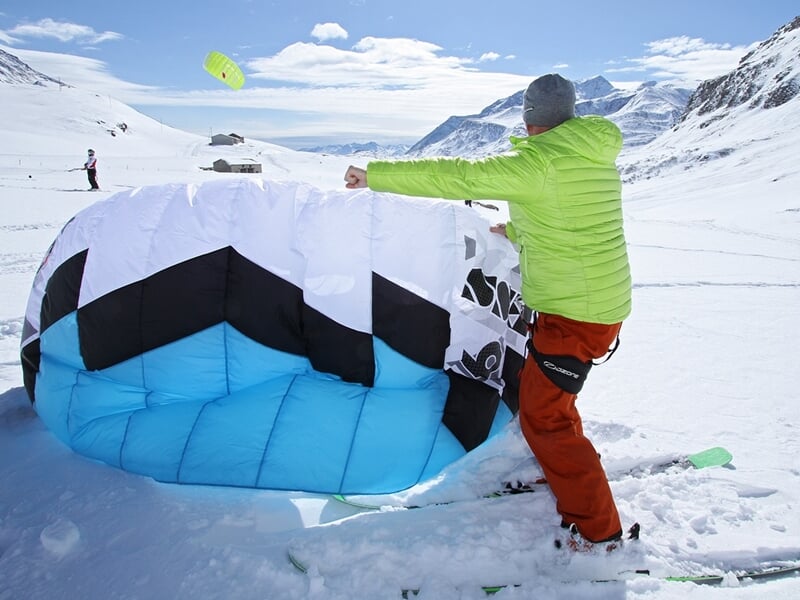 Un homme en veste verte et pantalon orange déploie une voile de snowkite sur la neige sous un ciel partiellement nuageux.