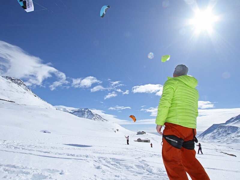 Un homme en veste verte regarde plusieurs voiles de snowkite colorées voler dans le ciel bleu au-dessus d'un paysage enneigé.