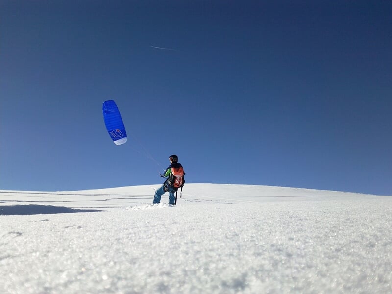 Un pratiquant de snowkite en tenue bleue et noire se tient sur la neige avec une voile bleue dans un ciel dégagé et lumineux.