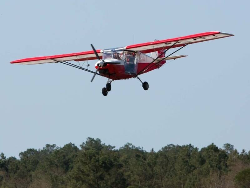 Avion ultraléger Skyranger rouge et blanc en vol au-dessus d'une forêt dense sous un ciel clair.