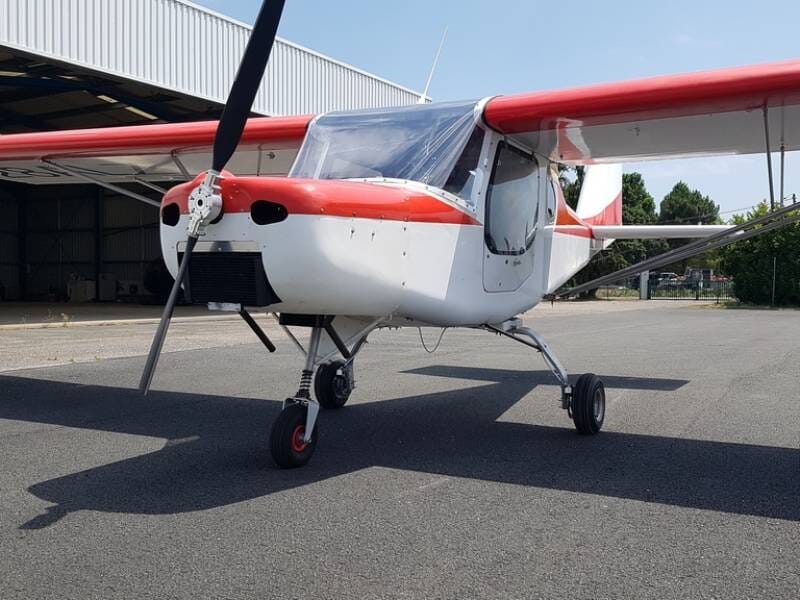 Avion ultraléger Buse Air 150 GT rouge et blanc stationné au sol devant un hangar sous un ciel dégagé.