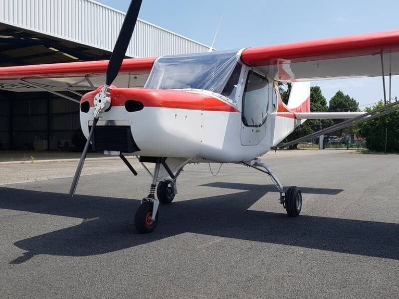 Avion ultraléger Buse Air 150 GT rouge et blanc stationné au sol devant un hangar sous un ciel dégagé.