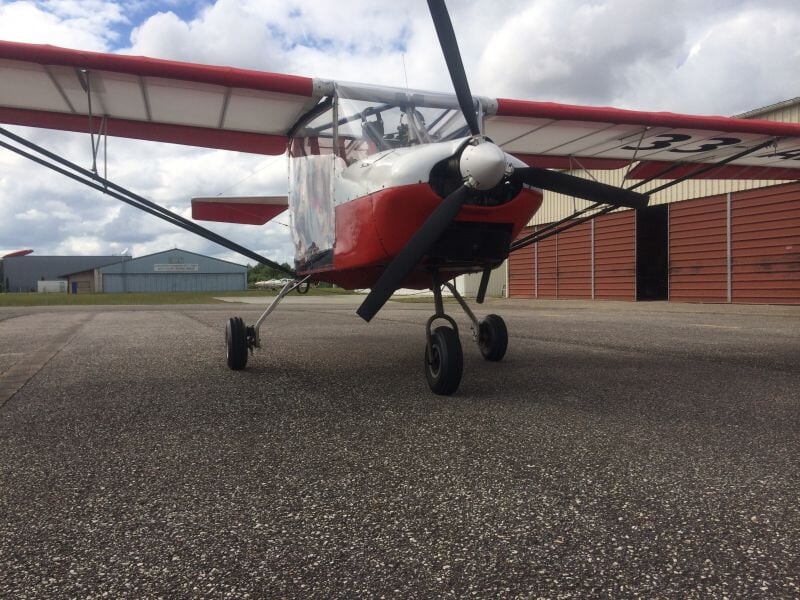 Avion ultraléger motorisé rouge et blanc stationné sur une piste en béton devant un hangar.