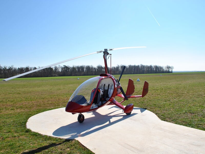 Gyrocoptère rouge stationné sur une piste en béton dans un champ vert sous un ciel bleu clair sans nuages.