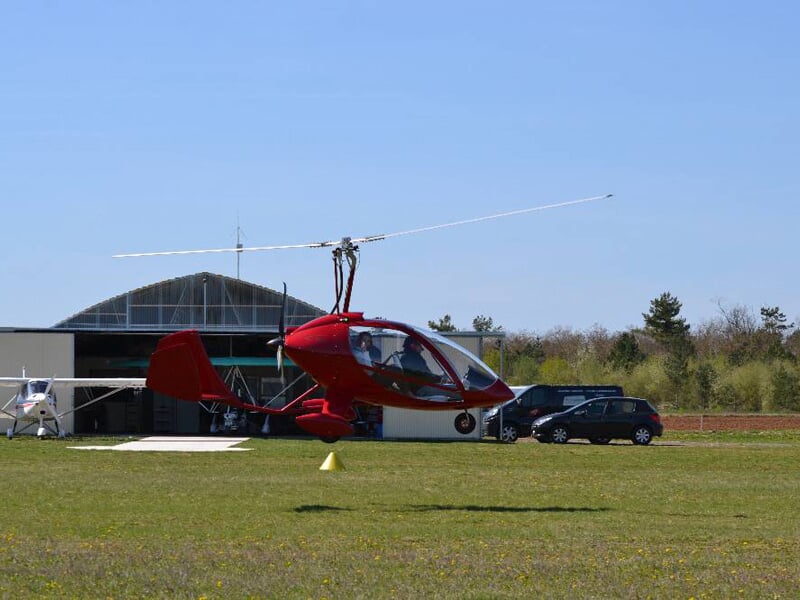 Gyrocoptère rouge en vol bas au-dessus d'un terrain herbeux près d'un hangar et de plusieurs voitures.