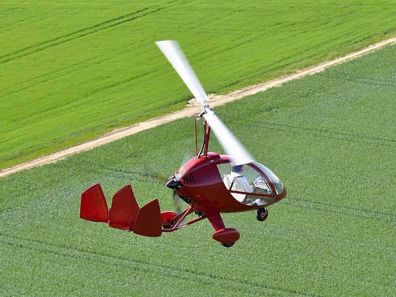 Gyrocoptère rouge en vol au-dessus d'un champ vert avec une route en terre visible à côté.