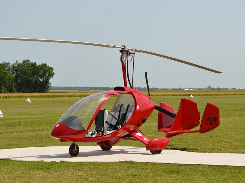Gyrocoptère rouge garé sur une piste en béton dans un champ vert avec un ciel bleu clair.