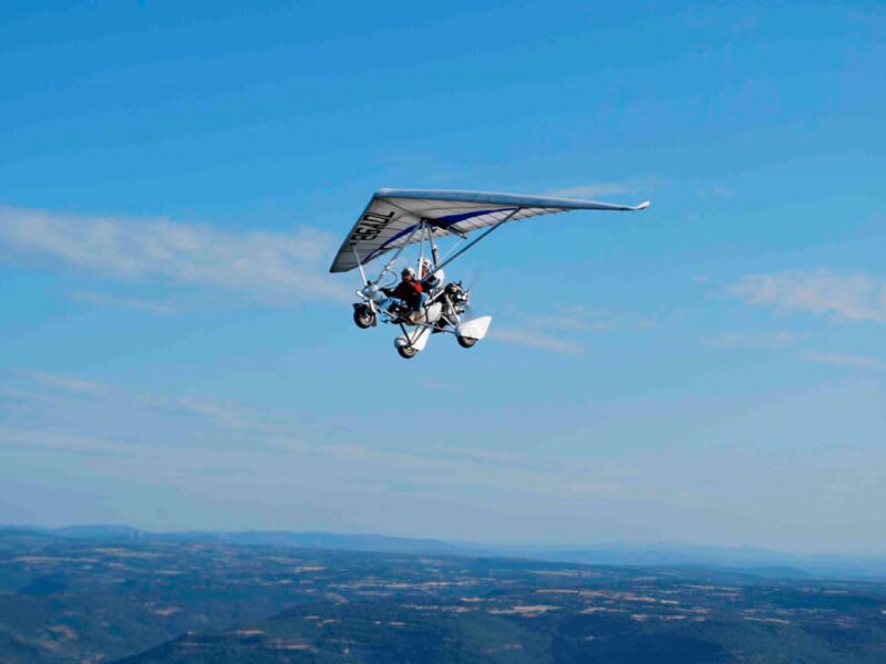ULM pendulaire en vol avec un pilote visible, survolant un paysage vallonné sous un ciel bleu clair.