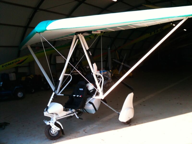 ULM pendulaire blanc stationné dans un hangar avec un sol en béton et un éclairage intérieur.