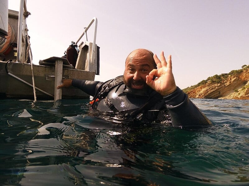 Plongeur barbu en combinaison noire sortant de l'eau, faisant un signe OK avec la main devant un bateau.