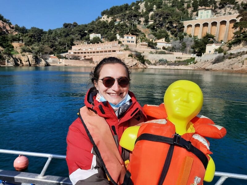 Femme souriante portant un gilet de sauvetage rouge, tenant une poupée de sauvetage jaune sur un bateau.