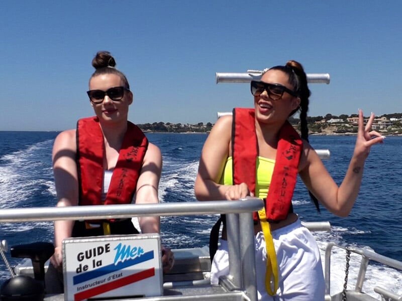 Deux femmes en gilets de sauvetage rouges sur un bateau, l'une fait un signe de paix avec la main.