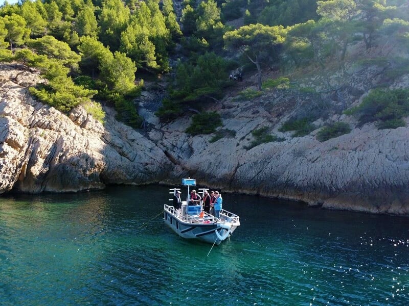 Petit bateau blanc avec plusieurs personnes à bord naviguant dans une crique entourée de falaises et de pins.