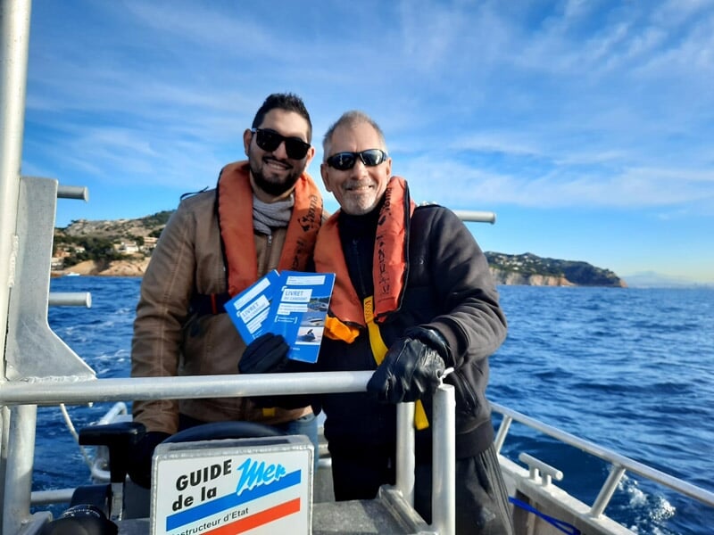 Deux hommes souriants en gilets de sauvetage sur un bateau, tenant des brochures bleues avec un panneau Guide de la Mer.