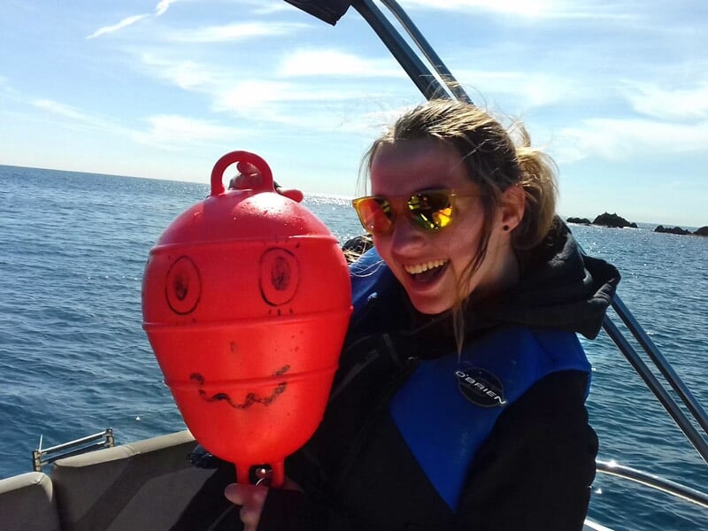 Femme souriante sur un bateau tenant une bouée rouge avec un visage dessiné, en mer calme et ensoleillée.