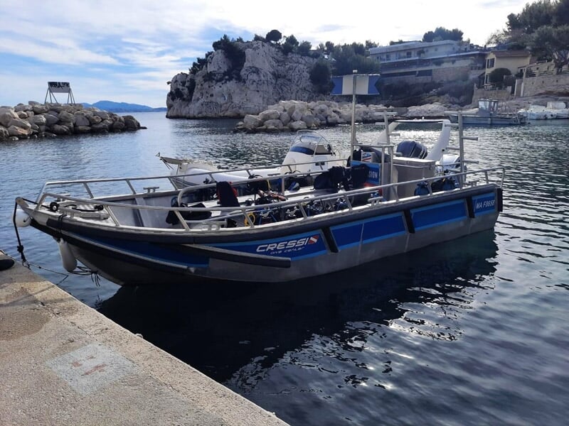 Petit bateau amarré au quai avec équipement de plongée visible, dans un port calme entouré de rochers.