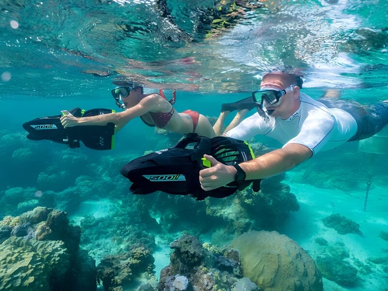 Deux plongeurs en mer avec palmes et scooter sous-marin, explorant un récif corallien sous l'eau claire.