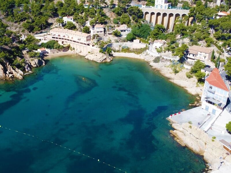 Vue aérienne d'une crique avec eau turquoise, rochers et maisons, idéale pour la plongée sous-marine.