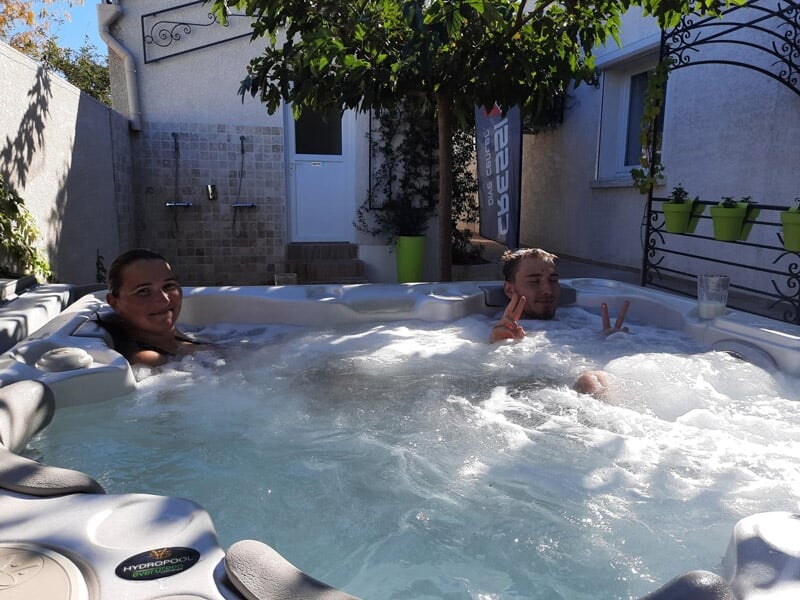 Deux hommes détendus dans un jacuzzi extérieur, entourés de murs blancs et d'un arbre avec des feuilles.