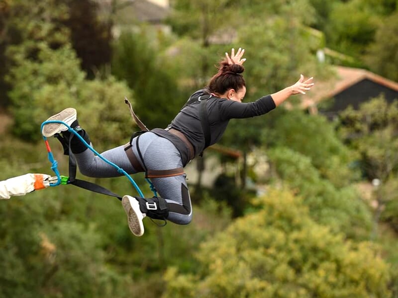 Femme effectuant un saut à l'élastique avec un harnais, entourée d'arbres et de maisons en arrière-plan.
