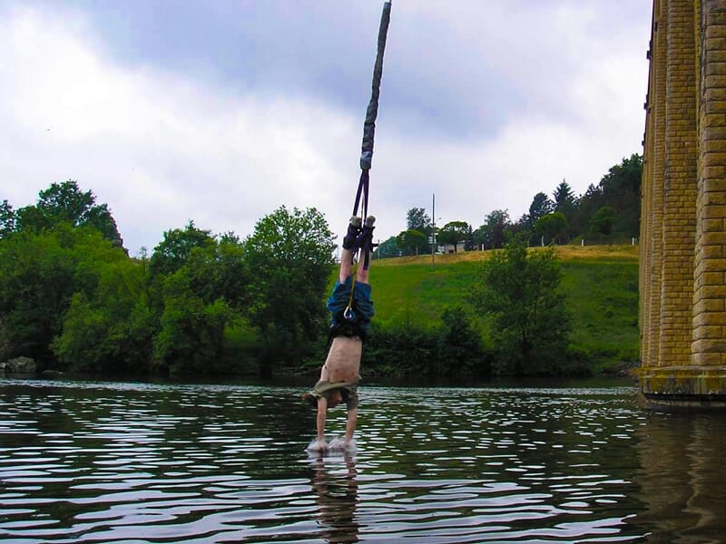 Personne suspendue la tête en bas lors d'un saut à l'élastique au-dessus d'une rivière avec un pont et des arbres en arrière-plan.