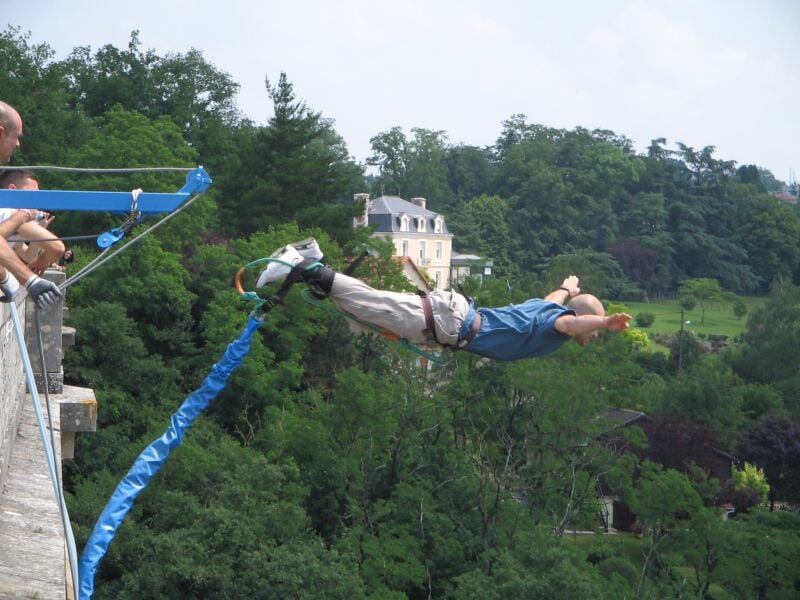 Homme effectuant un saut à l'élastique depuis un pont, entouré d'arbres et d'une maison en arrière-plan.