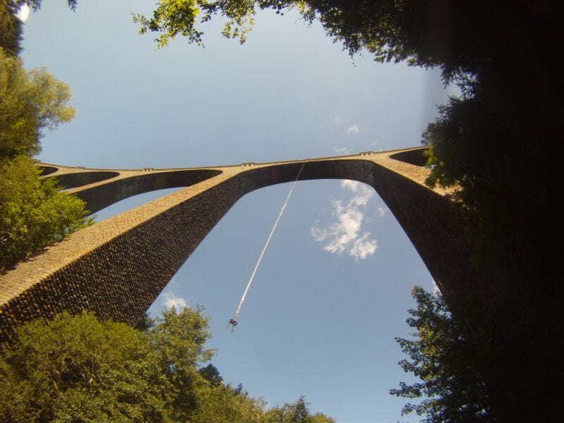 Personne effectuant un saut à l'élastique depuis un grand pont en pierre au-dessus d'une forêt dense sous un ciel bleu clair.