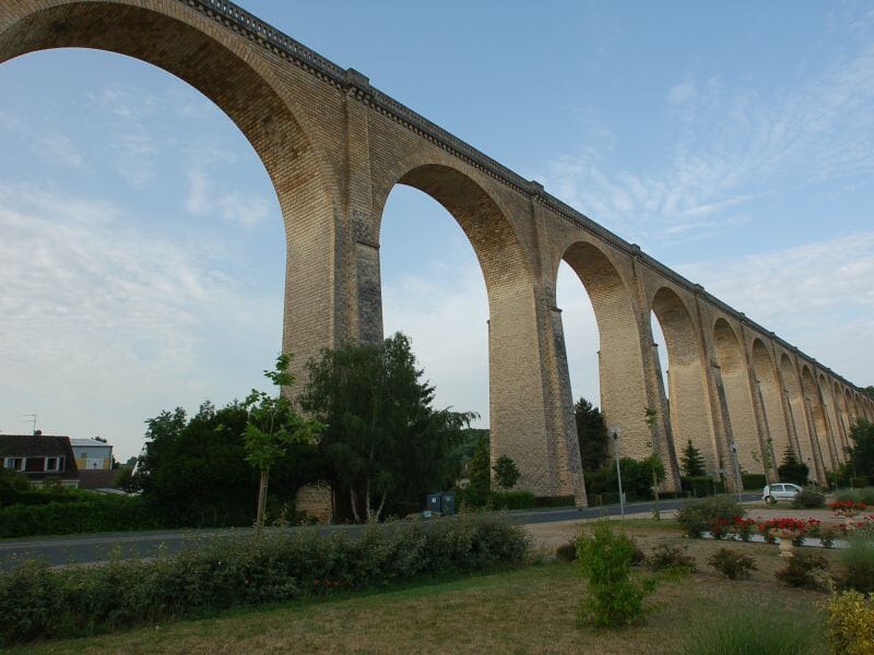 Viaduc de Le Blanc en pierre avec arches imposantes surplombant une route et un jardin fleuri sous un ciel clair.